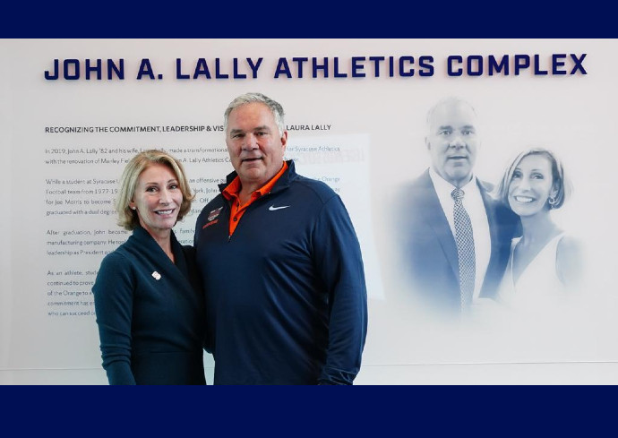 John and Laura Lally dressed in dark blue with Syracuse logo standing in front of wall announcing building of new athletics complex